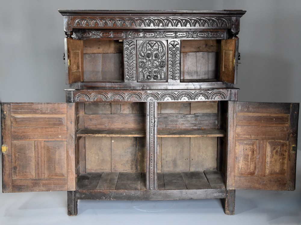 A 17th-century dark oak press cupboard featuring upper carved panel doors, overhanging canopy with pendant drops, and lower paneled double doors. View XI