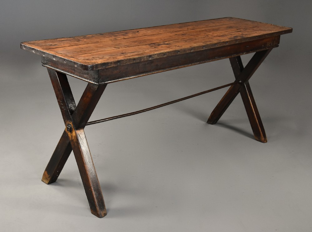 An early 19th-century sycamore tavern table featuring an X-frame trestle base, plank top, and metal stretcher rod.