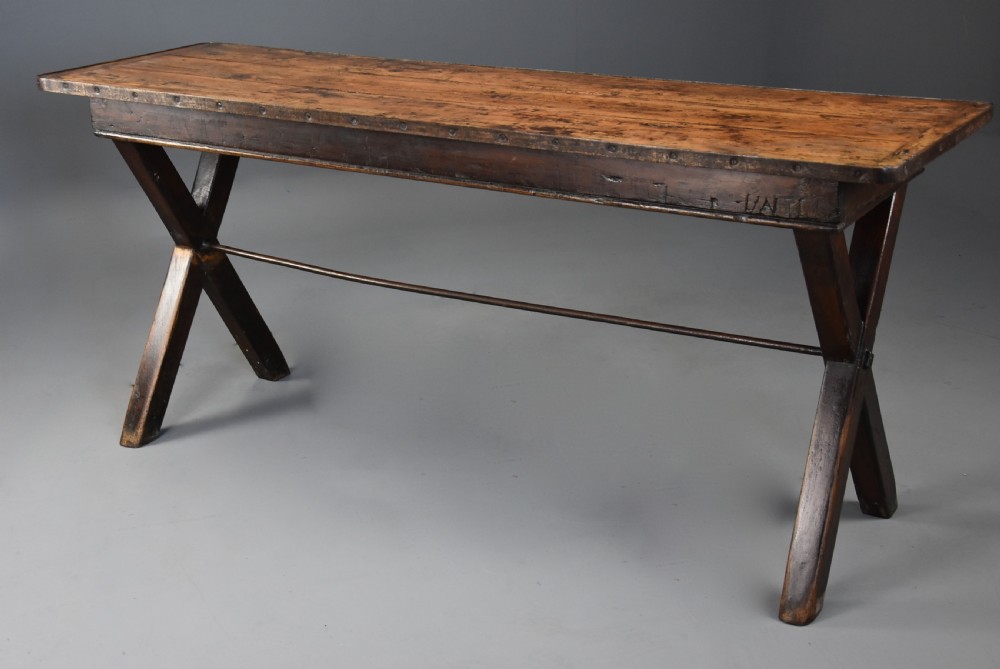 An early 19th-century sycamore tavern table featuring an X-frame trestle base, plank top, and metal stretcher rod. View X