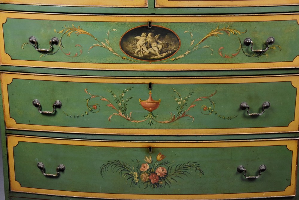 An early twentieth-century hand-painted green and gold bedroom suite featuring a wardrobe, chest of drawers with mirror, dressing desk, side tables, and caned chairs decorated with floral urn motifs. View X