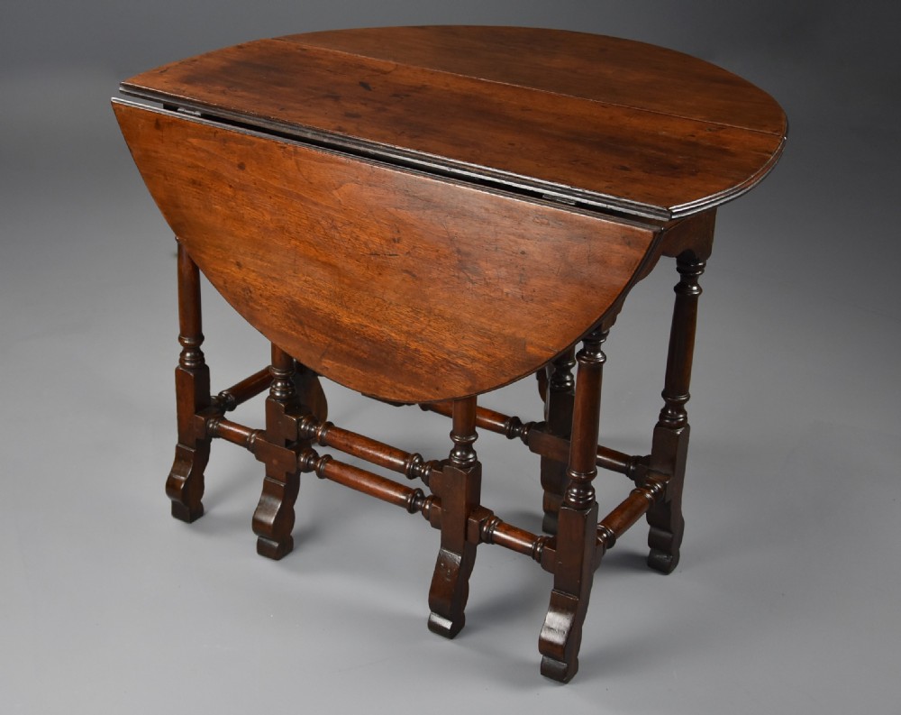 An 18th-century red walnut gateleg table featuring an oval top with two drop leaves, turned legs, box stretchers, and a small drawer with a brass knob. View IX