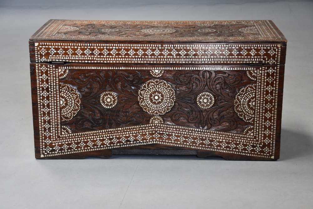 A dark hardwood rectangular storage trunk decorated with detailed relief carving and intricate geometric mother of pearl inlay across the front panel and lid. View X