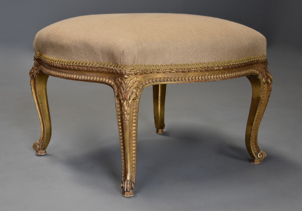 A mid-19th-century French carved giltwood footstool with a padded neutral linen seat, floral carving, and sweeping cabriole legs ending in scroll feet.