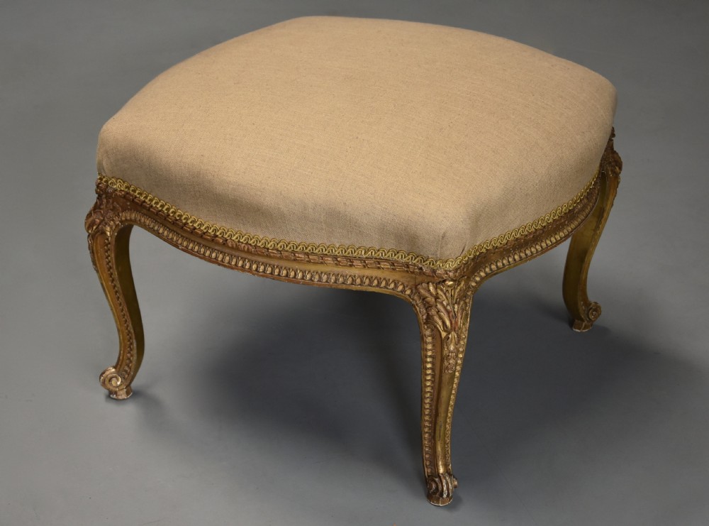 A mid-19th-century French carved giltwood footstool with a padded neutral linen seat, floral carving, and sweeping cabriole legs ending in scroll feet. View IV