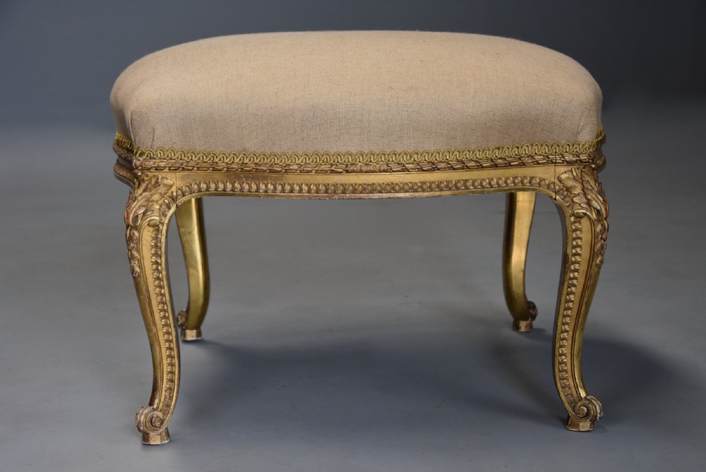 A mid-19th-century French carved giltwood footstool with a padded neutral linen seat, floral carving, and sweeping cabriole legs ending in scroll feet. View V