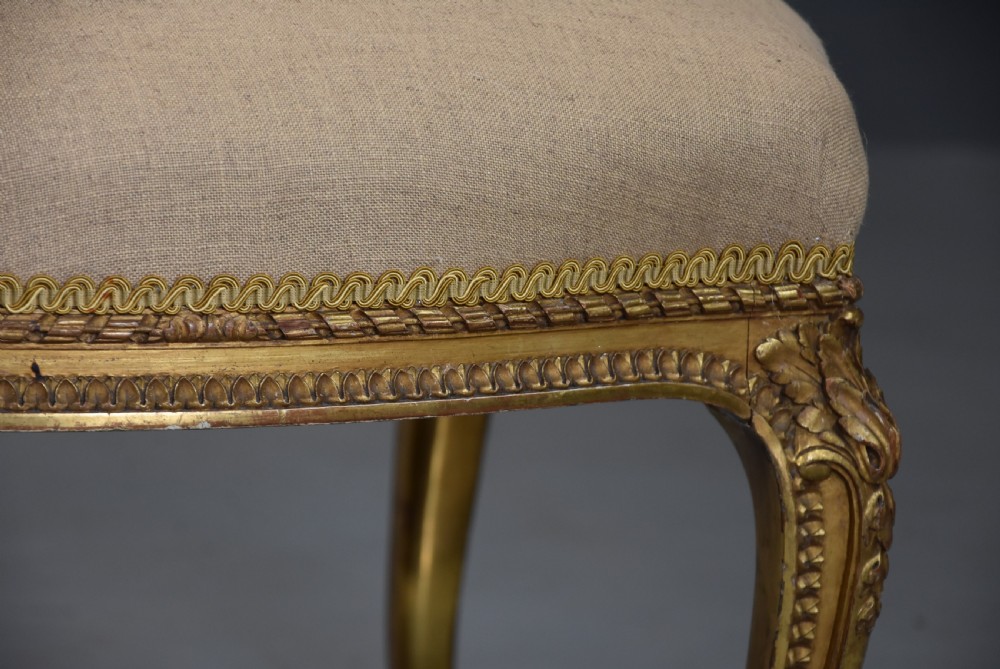 A mid-19th-century French carved giltwood footstool with a padded neutral linen seat, floral carving, and sweeping cabriole legs ending in scroll feet. View VI