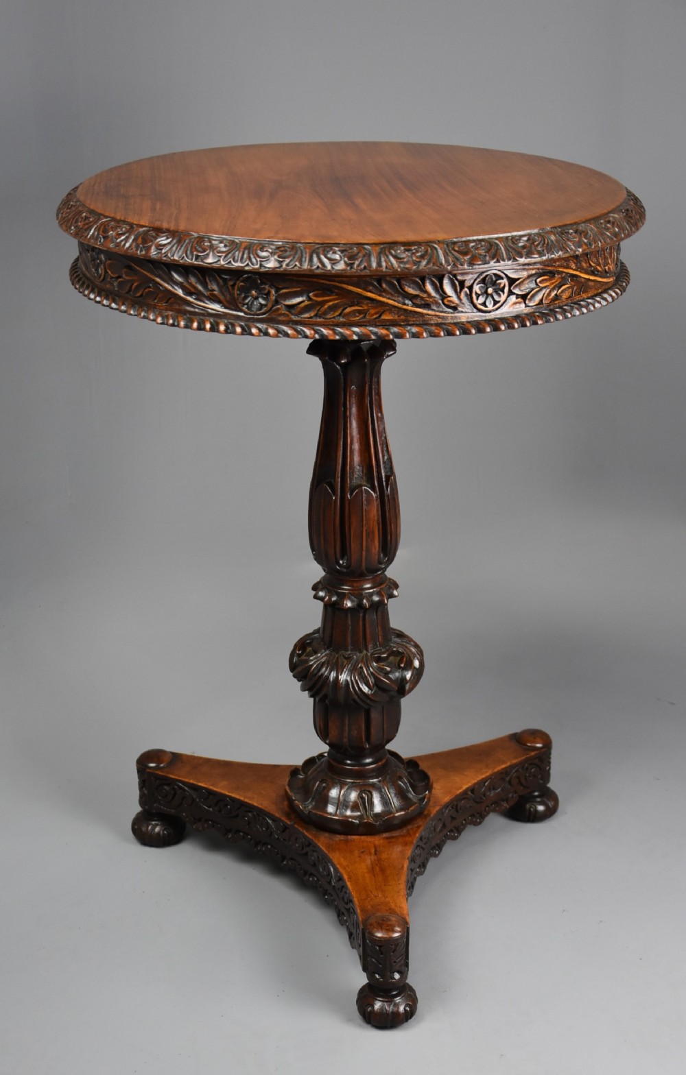 A mid-nineteenth-century Anglo-Indian circular padouk tilt-top occasional table featuring relief foliate carving along the apron, a turned pedestal, and a tripartite base.