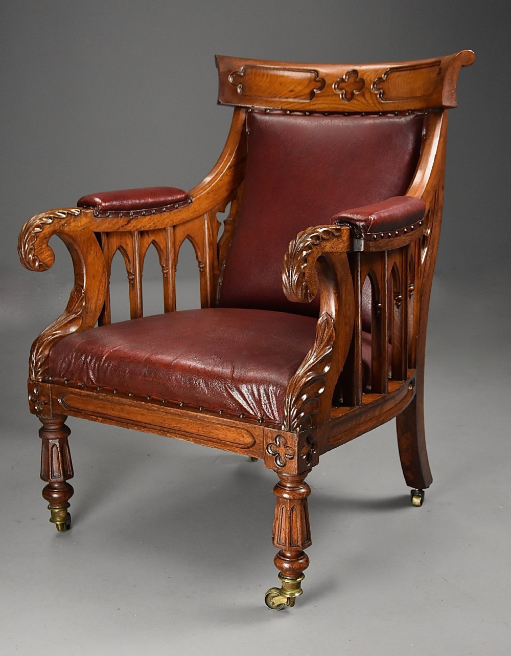 A nineteenth-century Gothic Revival carved oak armchair featuring deep burgundy leather upholstery, gothic tracery side panels, and turned legs on brass castors.