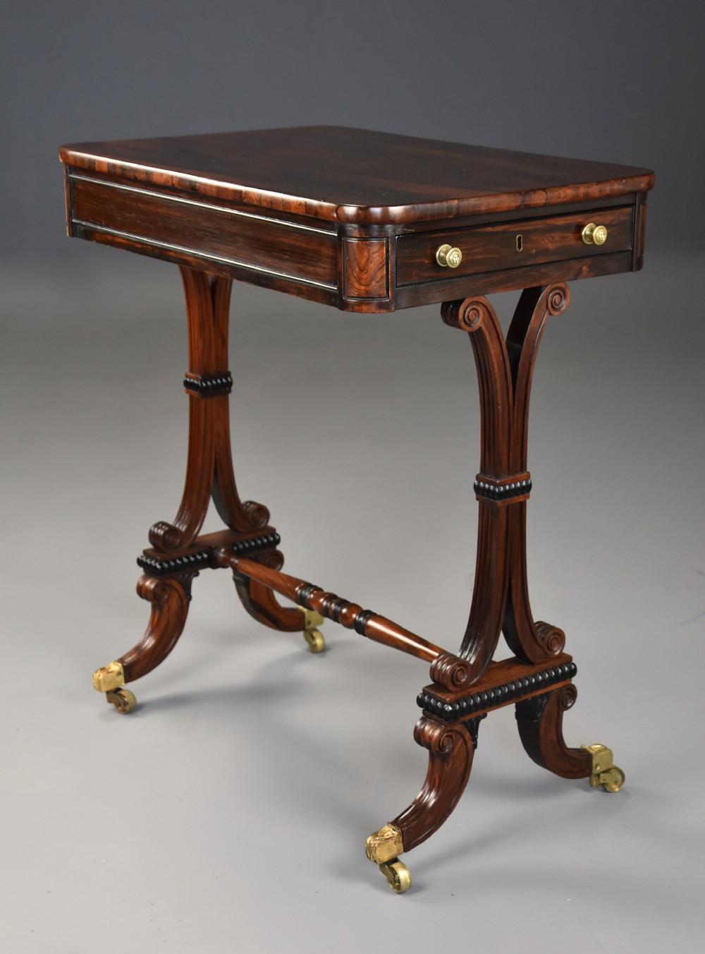 An early nineteenth-century rosewood occasional table featuring a rectangular top with a single drawer, scrolled trestle supports, beaded mouldings, and brass castors.