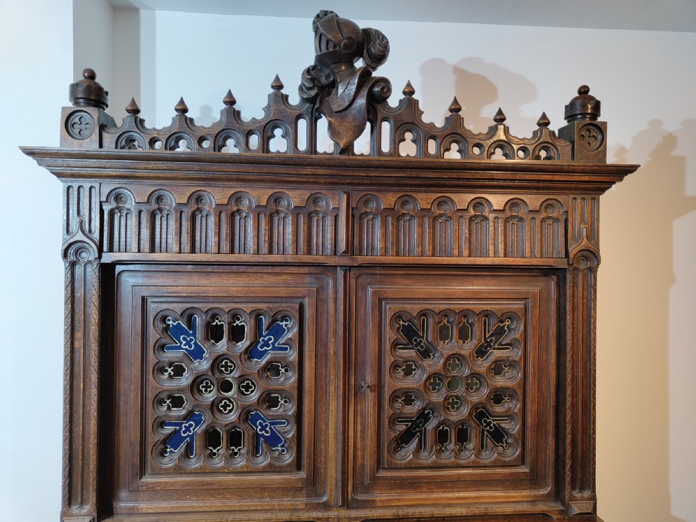 A two-tier Gothic Revival dark oak cabinet featuring carved architectural tracery, blue stained glass panel inserts, and profile portrait medallions on the lower doors. View III