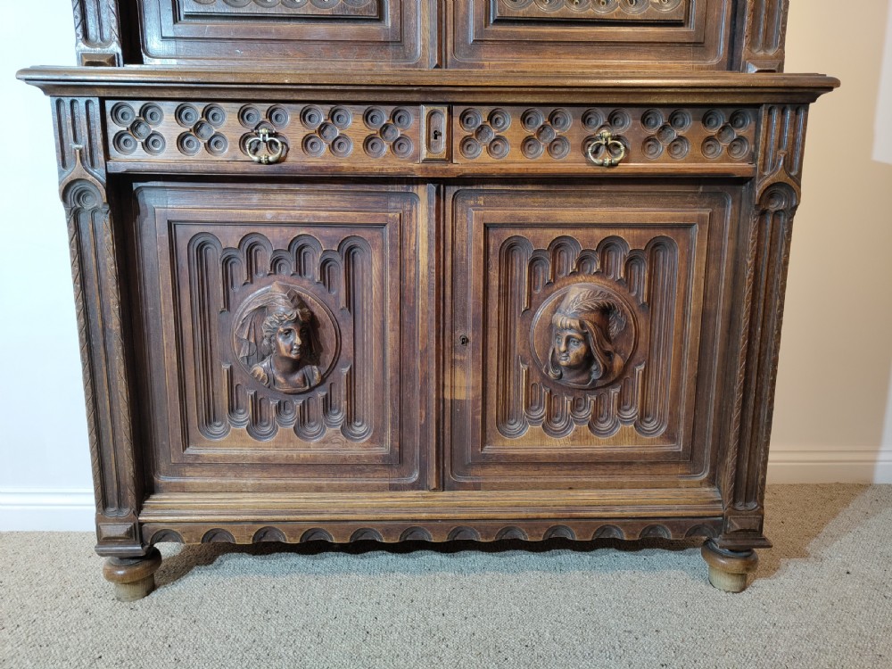 A two-tier Gothic Revival dark oak cabinet featuring carved architectural tracery, blue stained glass panel inserts, and profile portrait medallions on the lower doors. View IV