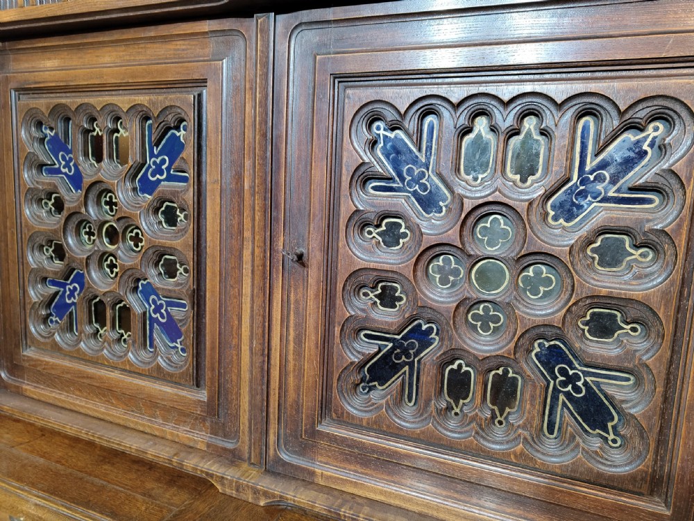 A two-tier Gothic Revival dark oak cabinet featuring carved architectural tracery, blue stained glass panel inserts, and profile portrait medallions on the lower doors. View VIII