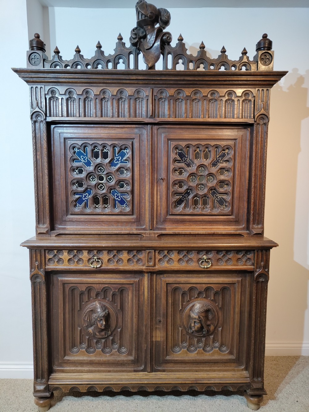 A two-tier Gothic Revival dark oak cabinet featuring carved architectural tracery, blue stained glass panel inserts, and profile portrait medallions on the lower doors.