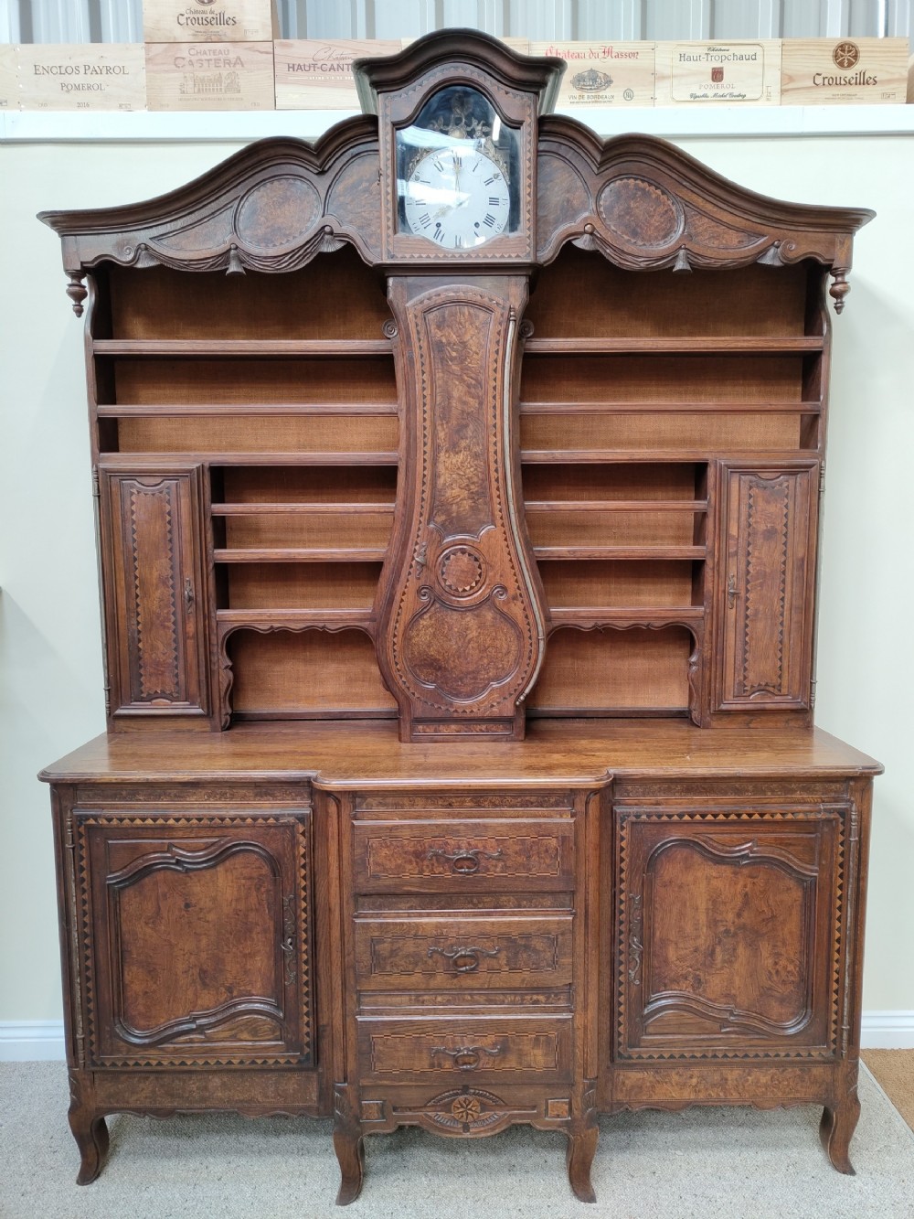 A carved oak French provincial clock dresser featuring an integrated central longcase clock, open shelving, and a lower sideboard with drawers and cupboards.