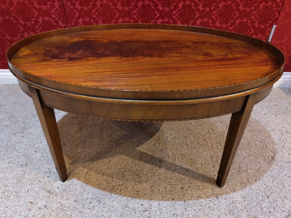 An oval mahogany coffee table with a raised tray edge detail, flame mahogany top veneer, and four square tapered legs.