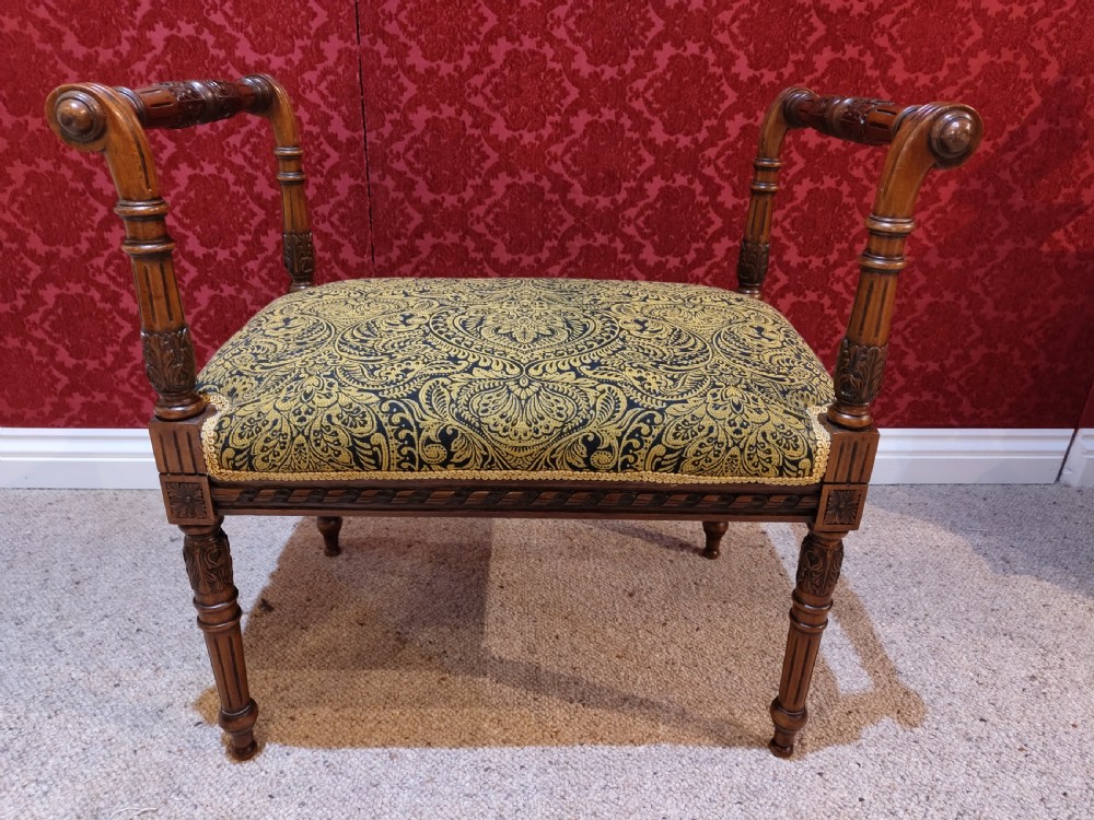 An antique carved wooden piano stool featuring raised side handles and a gold and black patterned upholstered cushion.