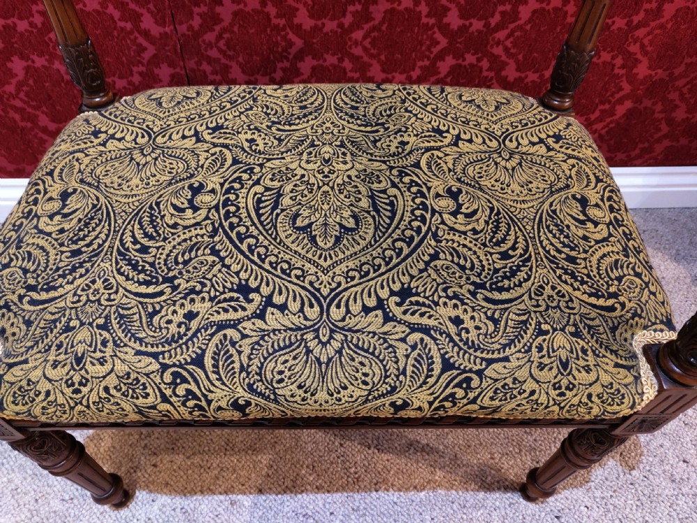 An antique carved wooden piano stool featuring raised side handles and a gold and black patterned upholstered cushion. View II
