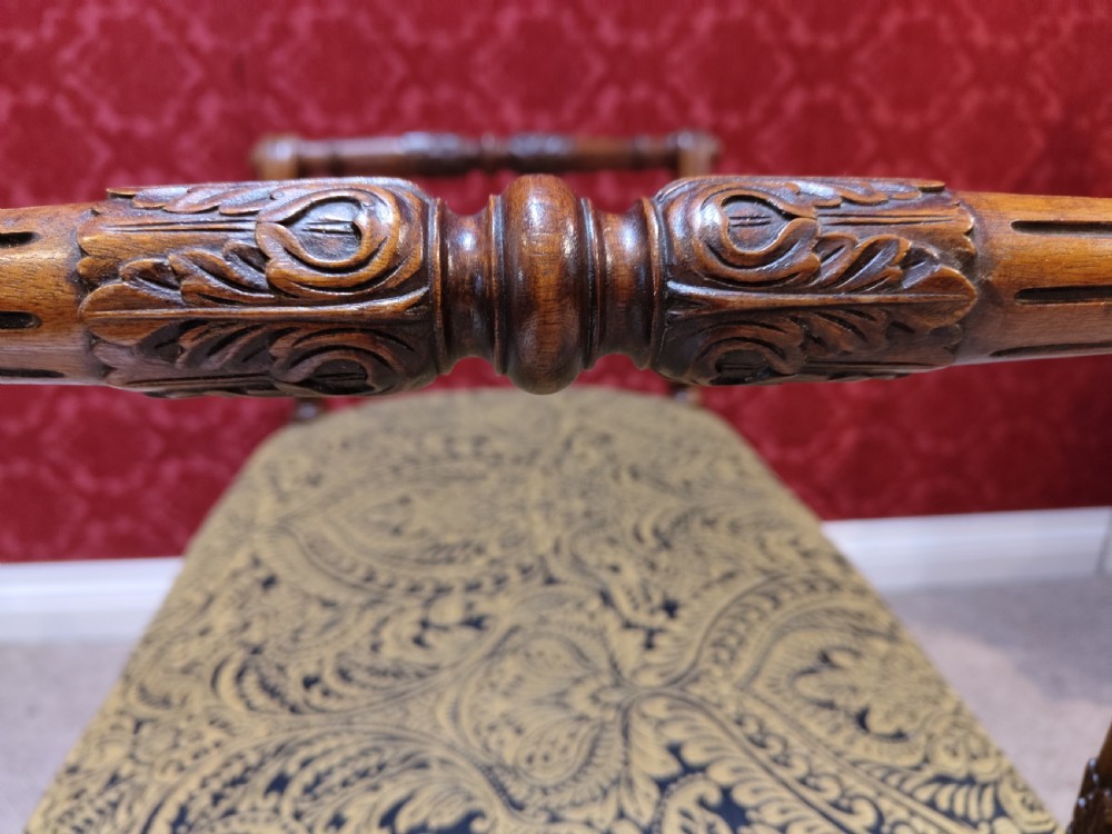 An antique carved wooden piano stool featuring raised side handles and a gold and black patterned upholstered cushion. View V