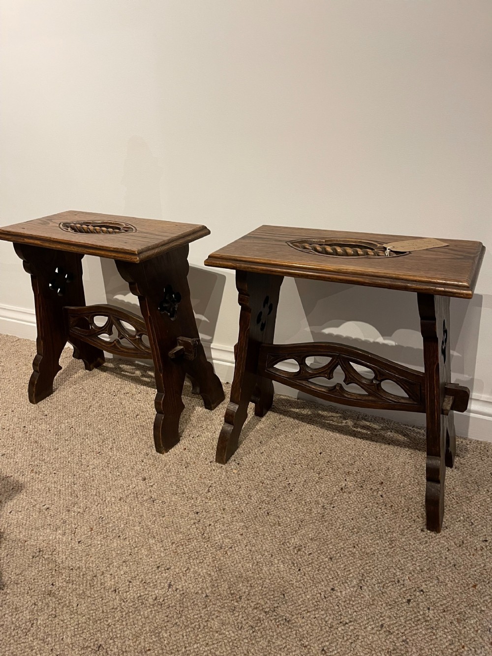 A pair of dark oak stools featuring carved Gothic tracery stretchers, trefoil side cut-outs, and integrated central seat handles.