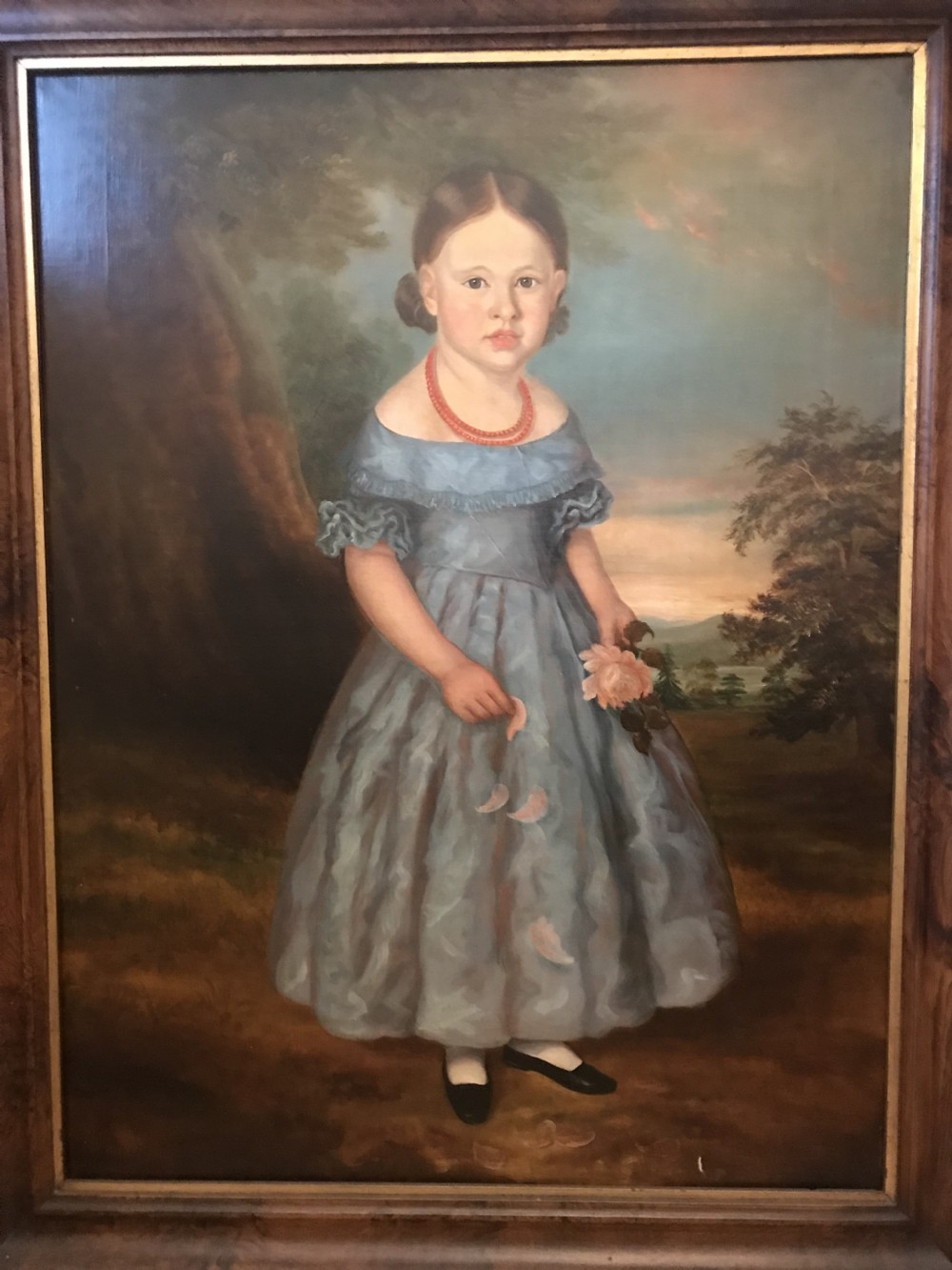 An antique oil painting depicting a standing young girl in a blue dress holding a rose, set within a wide timber frame with a gilded inner rim. View XIV