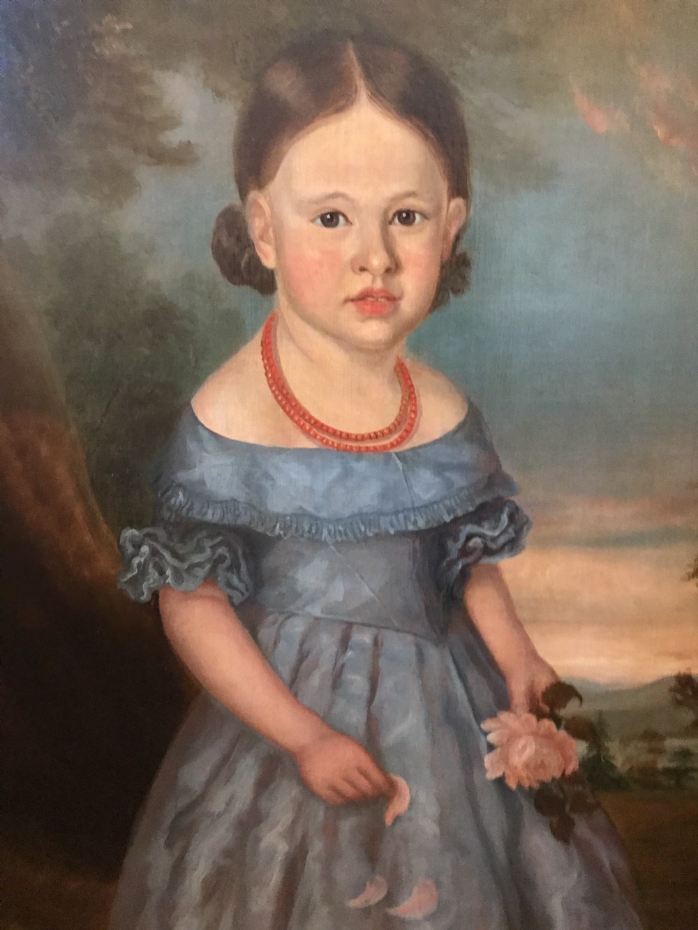 An antique oil painting depicting a standing young girl in a blue dress holding a rose, set within a wide timber frame with a gilded inner rim. View XV