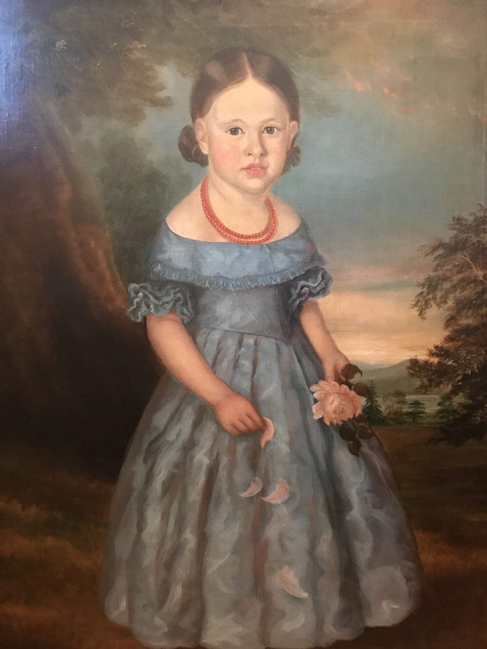 An antique oil painting depicting a standing young girl in a blue dress holding a rose, set within a wide timber frame with a gilded inner rim. View XVI