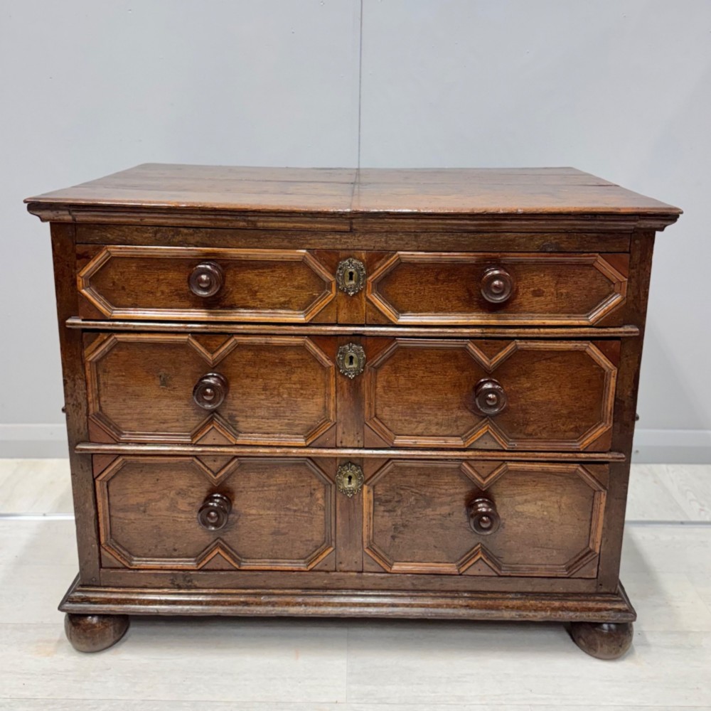 A seventeenth-century dark oak chest featuring three drawers decorated with geometric panel mouldings, wooden turned handles, brass escutcheons, and bun feet.