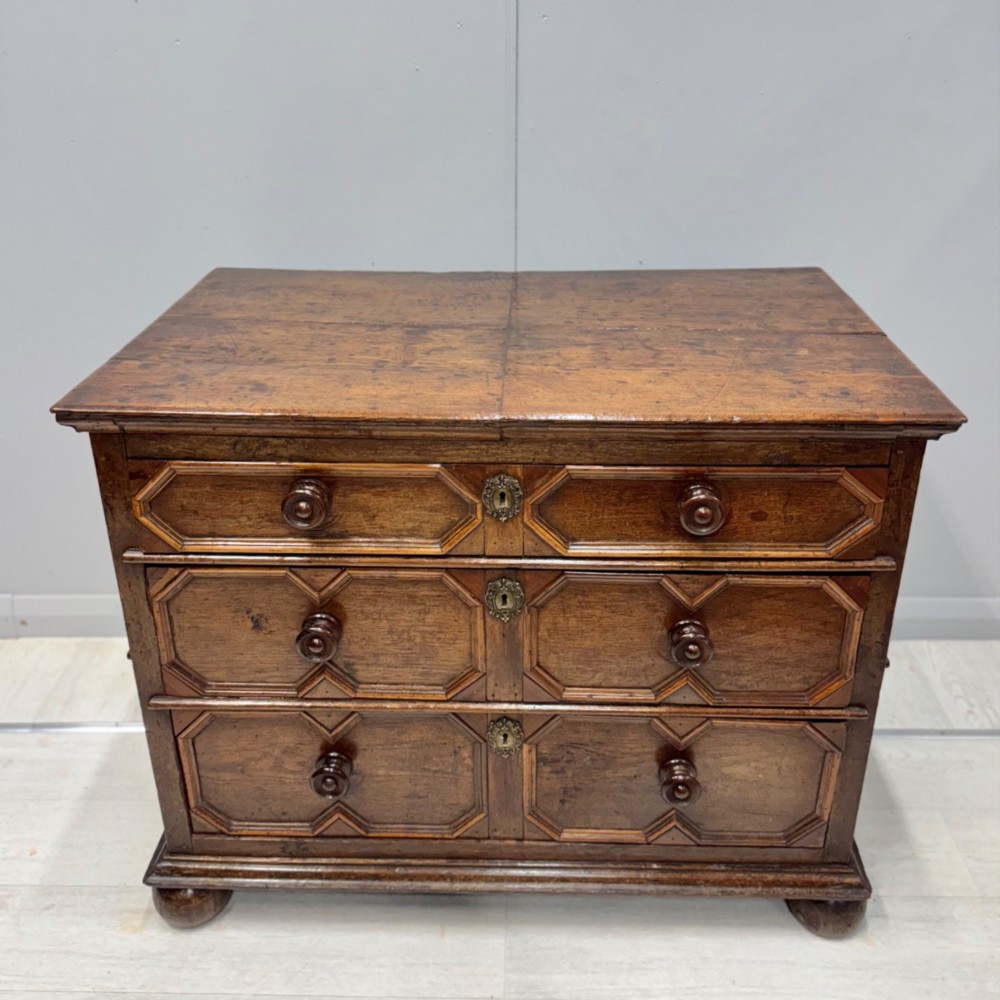A seventeenth-century dark oak chest featuring three drawers decorated with geometric panel mouldings, wooden turned handles, brass escutcheons, and bun feet. View V
