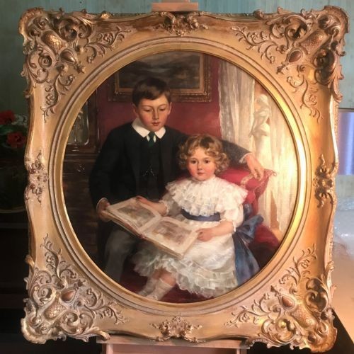 An Edwardian oil portrait painting depicting a young boy and girl reading a book together, presented within a square gilt frame featuring an oval aperture.