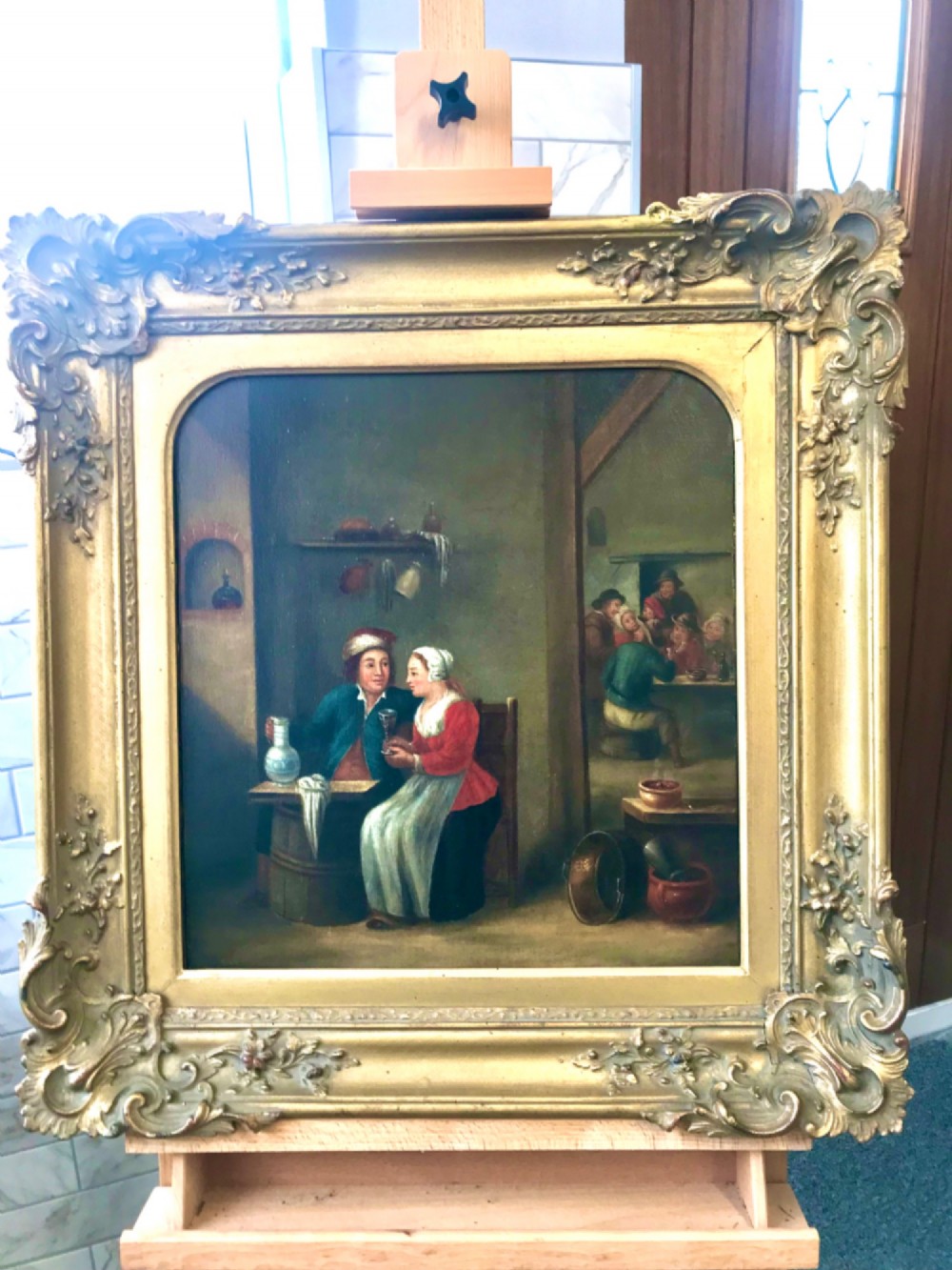 An 18th-century style oil painting depicting figures inside a tavern interior, set within a carved gold giltwood frame.
