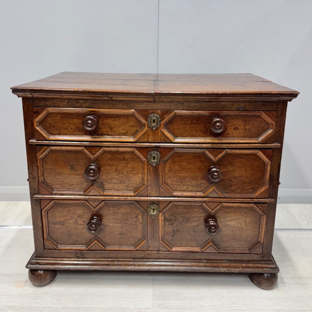 A 17th-century dark oak chest of drawers featuring geometric mitred mouldings on three drawer fronts, turned wooden handles, brass escutcheons, and bun feet.