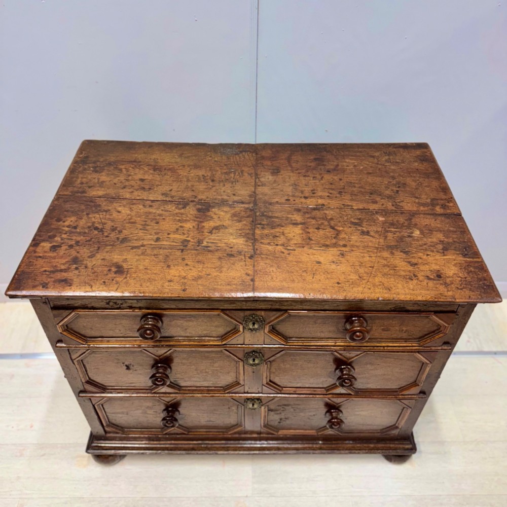 A 17th-century dark oak chest of drawers featuring geometric mitred mouldings on three drawer fronts, turned wooden handles, brass escutcheons, and bun feet. View XII