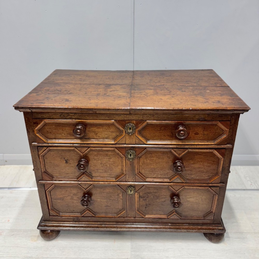 A 17th-century dark oak chest of drawers featuring geometric mitred mouldings on three drawer fronts, turned wooden handles, brass escutcheons, and bun feet. View XIII