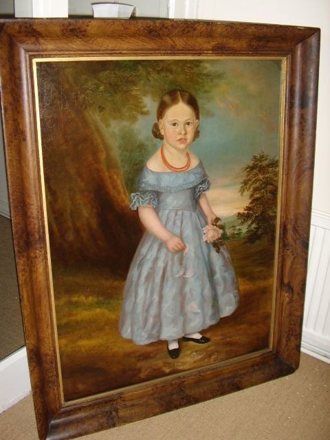 An antique oil portrait on canvas depicting a young girl in a blue dress holding a rose, displayed within a wide flame wood frame.