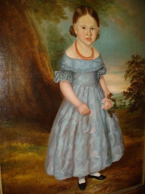 An antique oil portrait on canvas depicting a young girl in a blue dress holding a rose, displayed within a wide flame wood frame. View VI