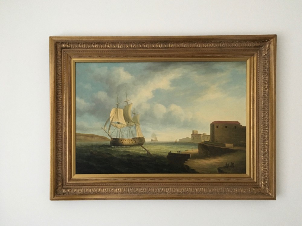 A 19th-century style marine oil painting on panel depicting a large sailing warship anchored near a stone coastal fortification under a cloudy sky, presented in a heavily moulded gold gilt frame. View XIII