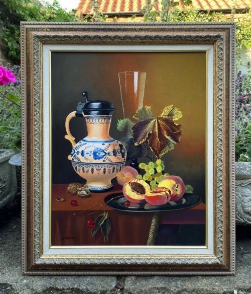 A framed oil painting depicting a still life composition with peaches, grapes, cherries, a decorated ceramic jug, and a glass vessel on a table against a warm brown background.