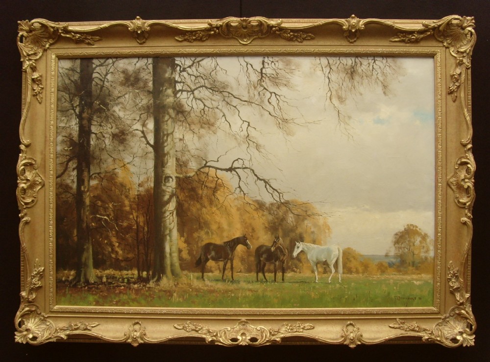 An oil painting in an ornate gilded frame depicting three horses standing in a green meadow backed by autumn trees.