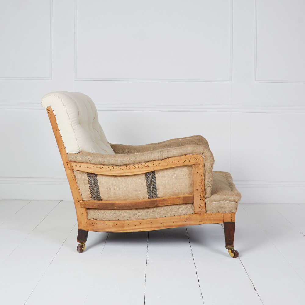 A deep-seated Howard style armchair shown in natural calico and hessian under-upholstery with an exposed wooden frame and brass castors.
