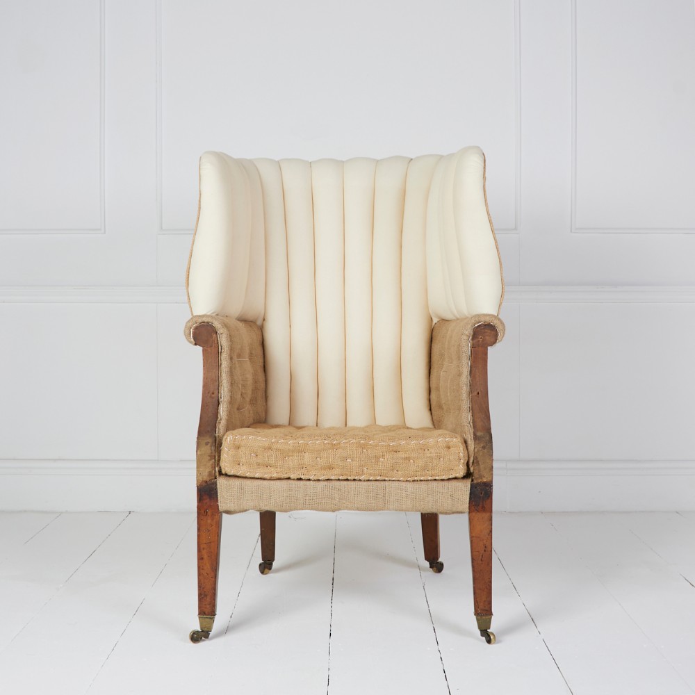 An antique barrel back wing armchair with vertical channel-tufted cream calico upholstery, hessian trim, and tapered mahogany legs on brass castors.