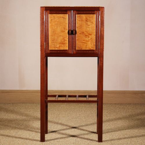fig 1. A tall mahogany cabinet on stand featuring contrasting light wood door panels with inlaid stringing borders and a slatted lower shelf.