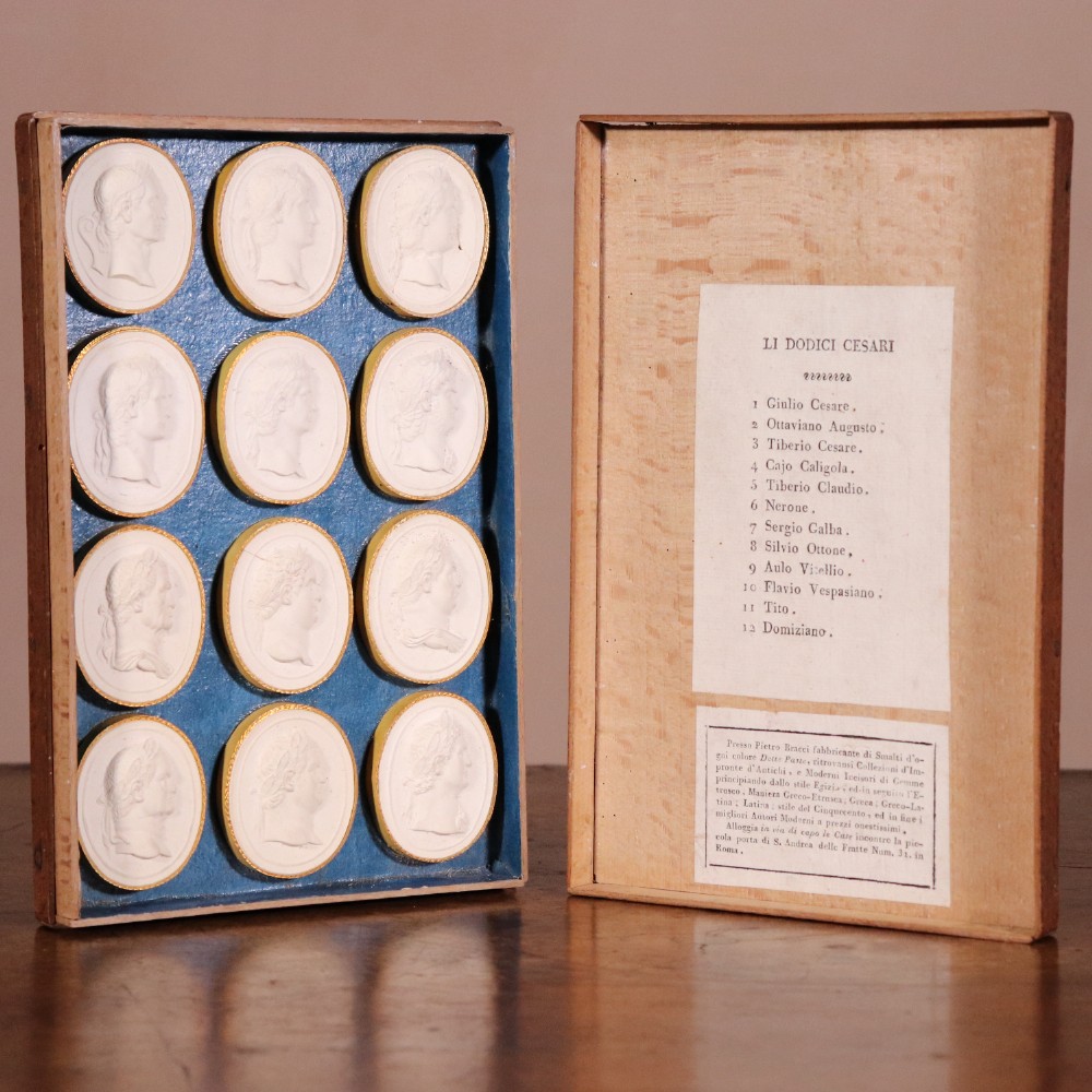 A set of twelve oval white plaster relief intaglios depicting Roman emperors mounted inside a tray with blue backing and printed Italian text labels.