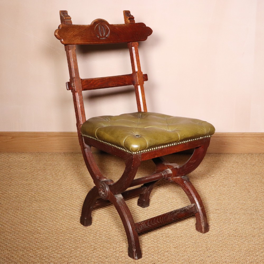 A 19th-century Gothic Revival oak side chair featuring an X-frame base, carved backrest motif, and an olive green button-tufted leather seat secured with brass studs.