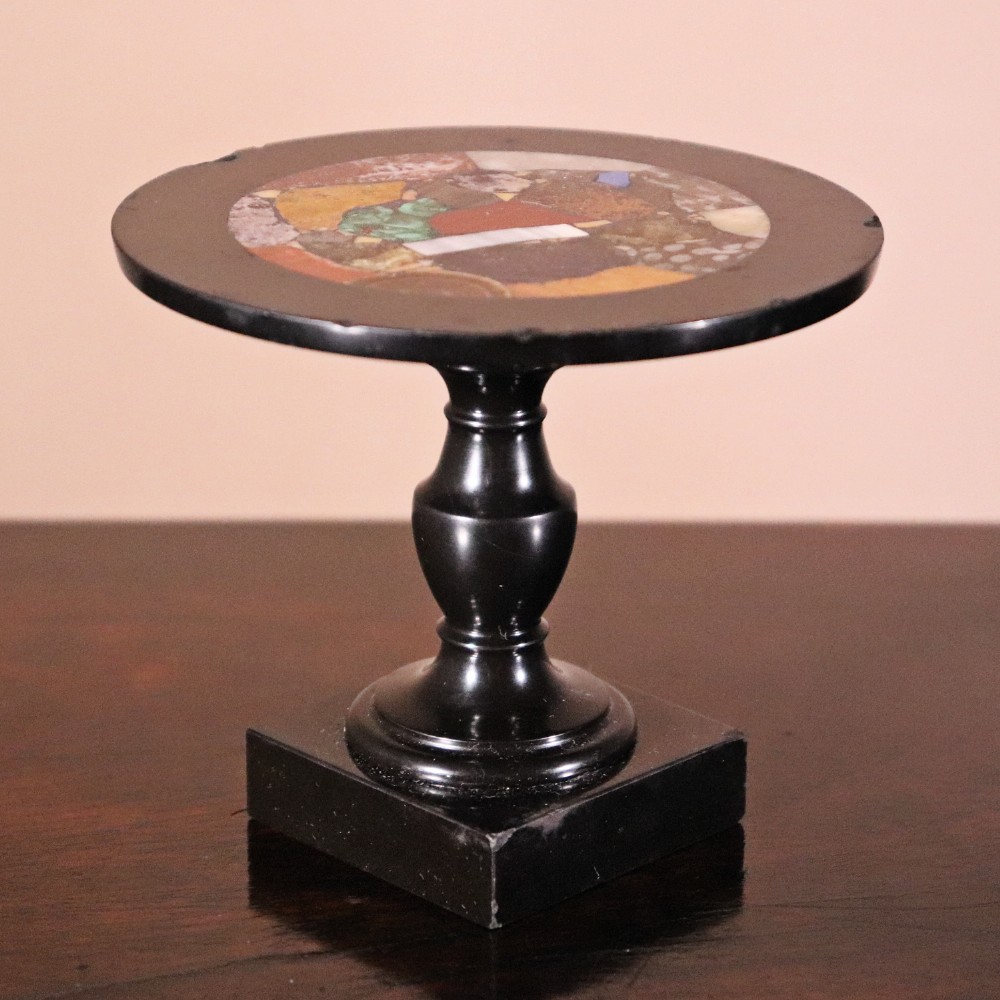A nineteenth-century Ashford black marble miniature table featuring a round top inlaid with colourful geometric marble specimens, supported by a turned baluster column on a square plinth base.