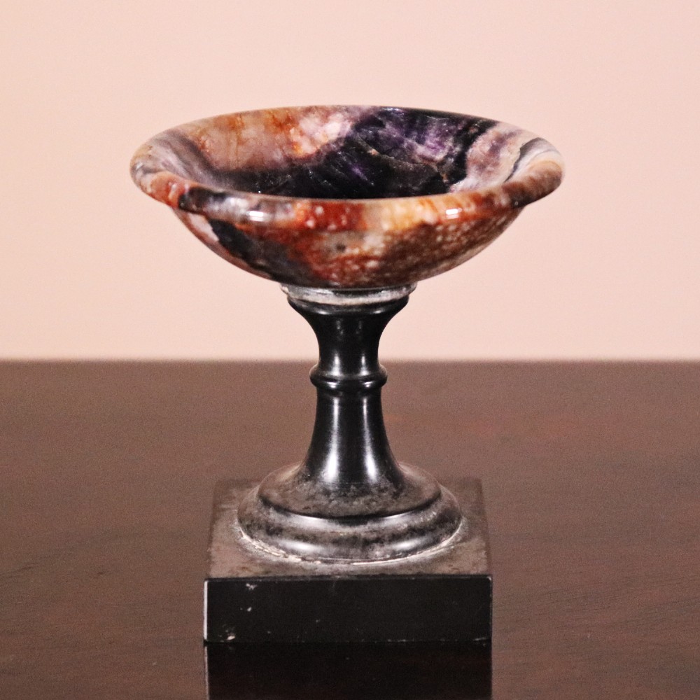 A small nineteenth-century tazza featuring a shallow turned Blue John bowl with purple and brown banding, set upon a black turned marble stem and square base.
