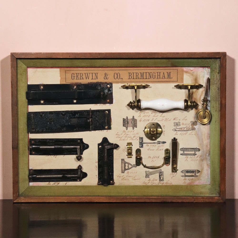 A framed commercial sample board containing antique brass and cast iron door hardware, latches, and ceramic handles mounted on annotated paper with a header reading Gerwin & Co., Birmingham.