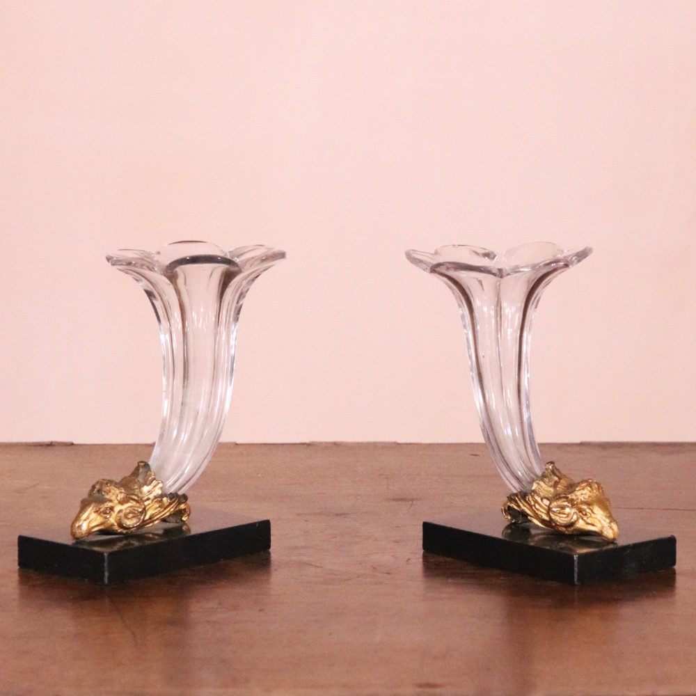 A pair of Regency cornucopia vases featuring fluted clear glass horns set into gilt bronze rams head mounts on rectangular black marble bases.