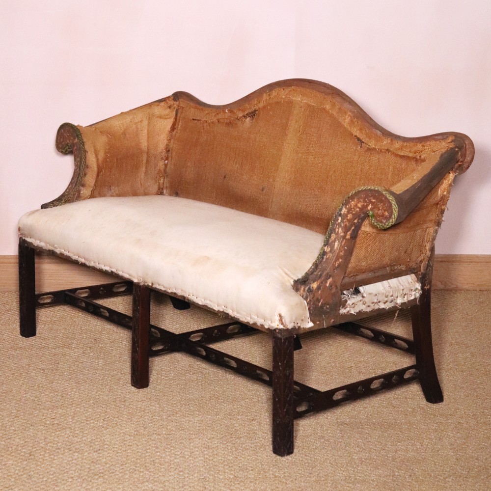 An antique Chippendale style mahogany settee featuring a serpentine camel back, scrolled arms, pierced stretchers, and exposed base lining fabric.