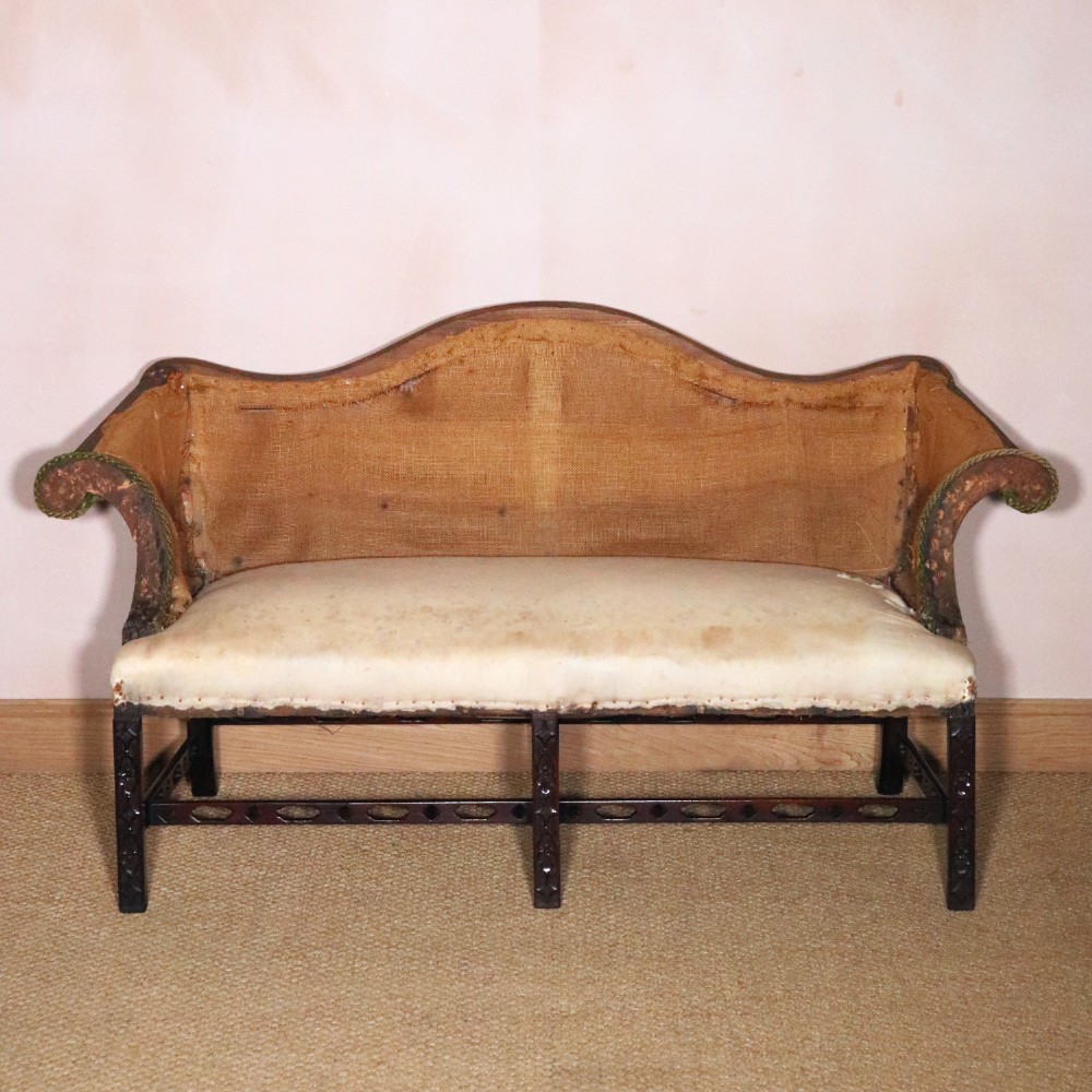 An antique Chippendale style mahogany settee featuring a serpentine camel back, scrolled arms, pierced stretchers, and exposed base lining fabric. View II