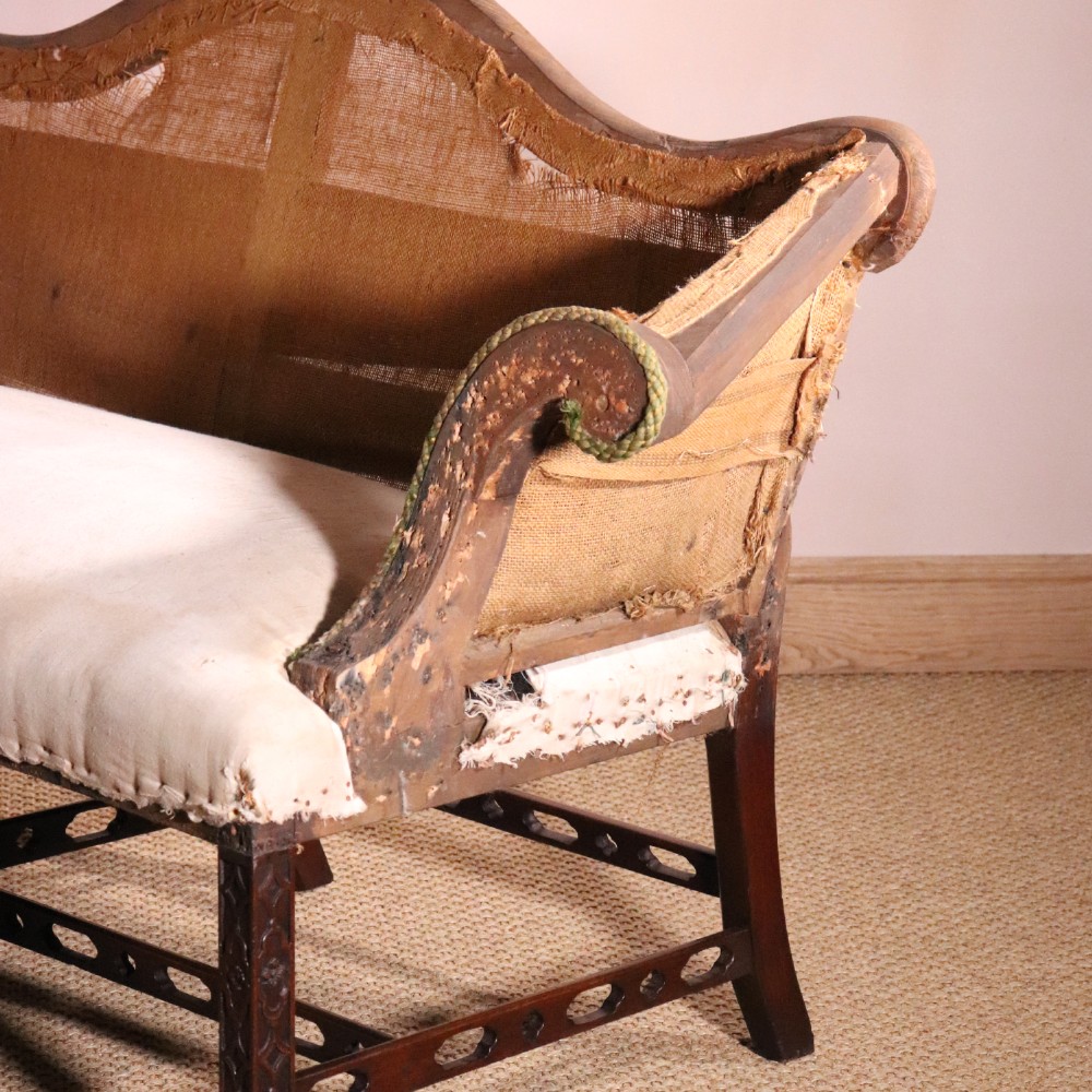 An antique Chippendale style mahogany settee featuring a serpentine camel back, scrolled arms, pierced stretchers, and exposed base lining fabric. View III